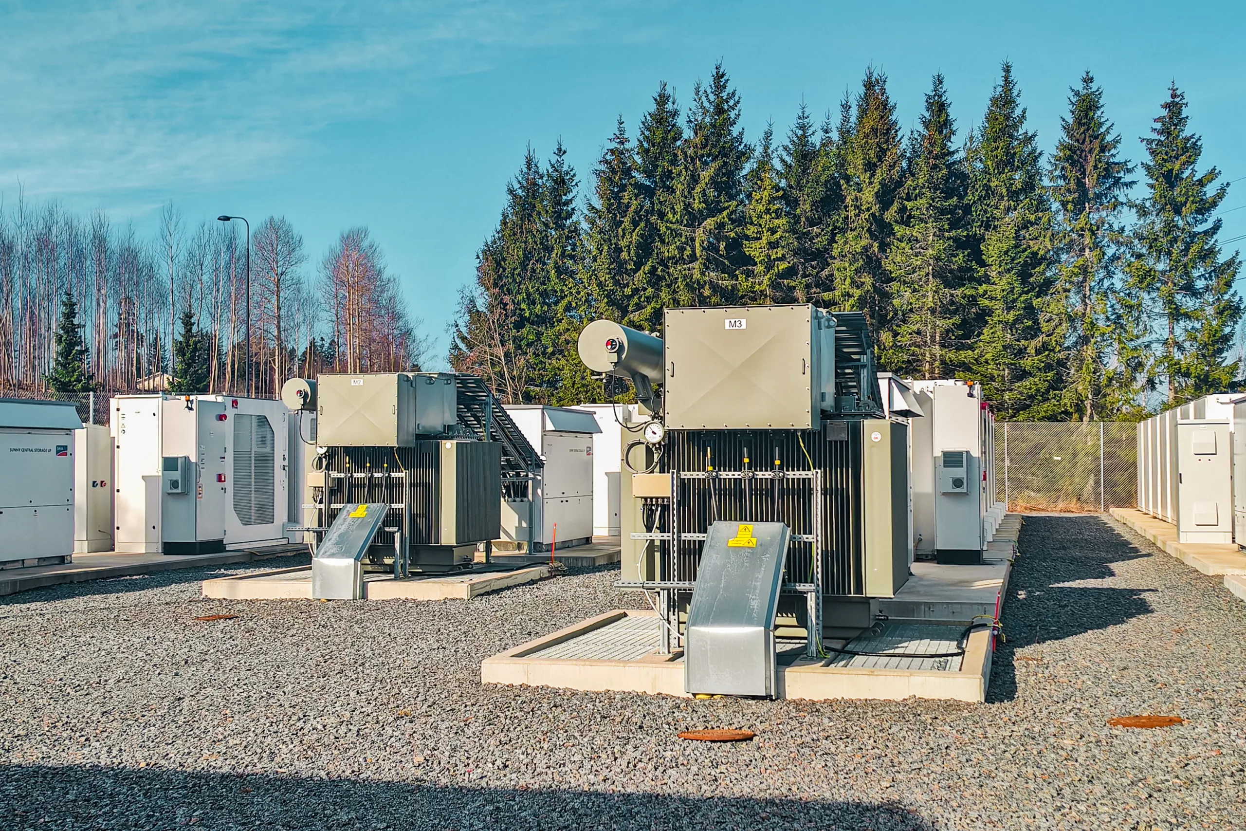 Ikaalinen battery storage facility showing transformer equipment, high-voltage power lines, and auxiliary buildings with forested backdrop under clear sky.