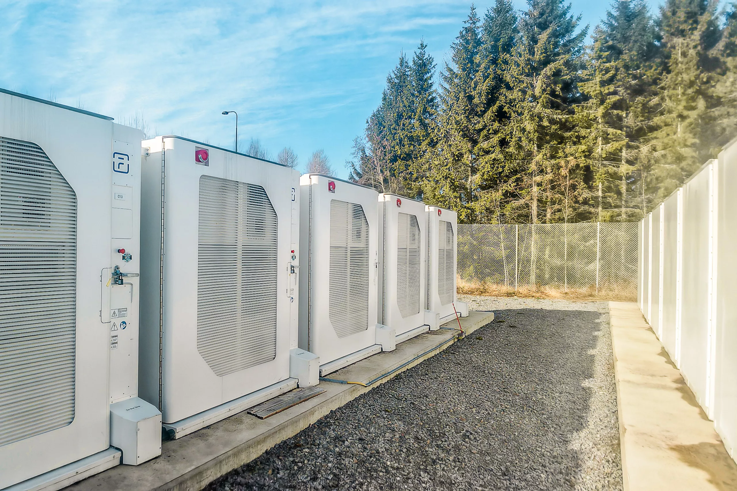 Modular battery storage units with transformer blocks and cooling components arranged on gravel surface with coniferous forest backdrop