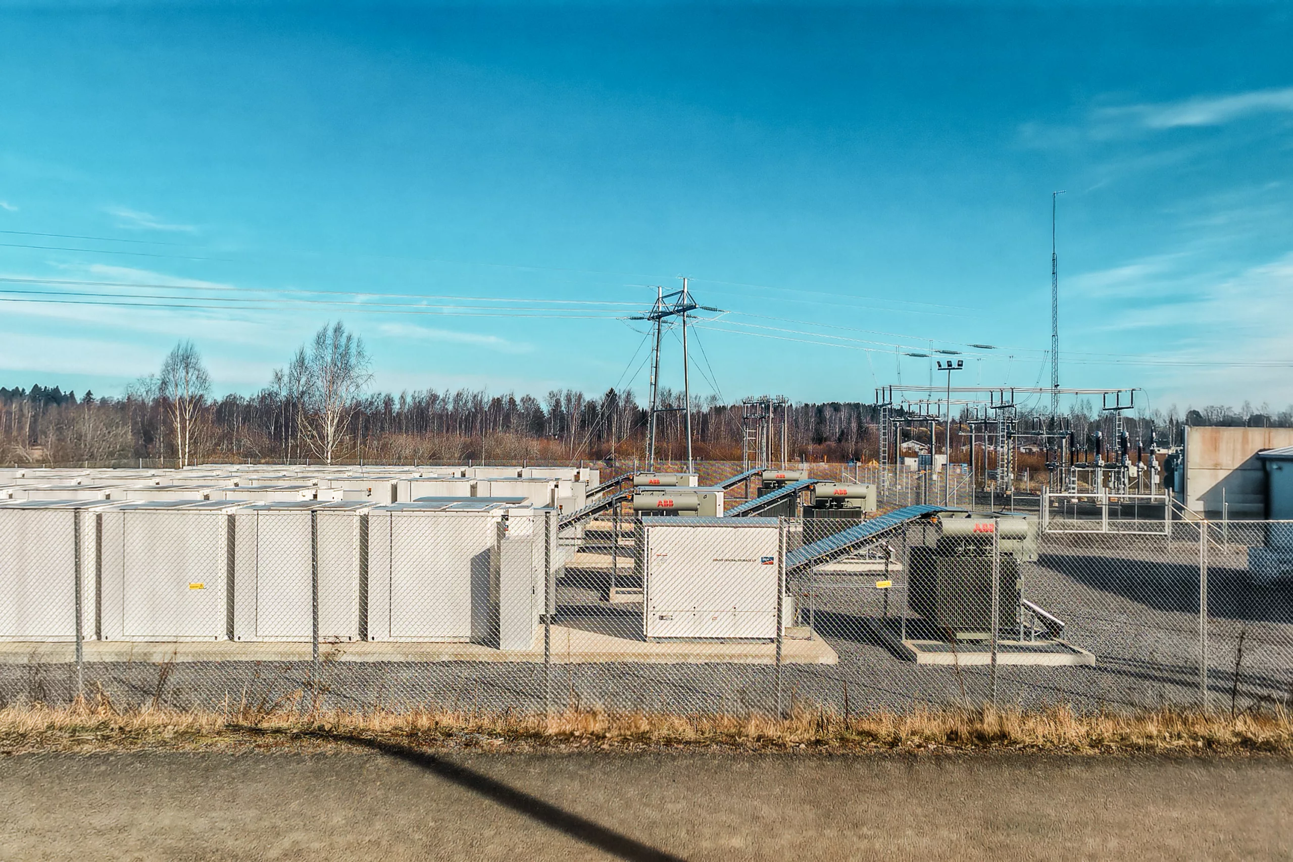 Series of white battery storage containers with cooling fins and red indicator lights, fenced enclosure in forested landscape under blue sky