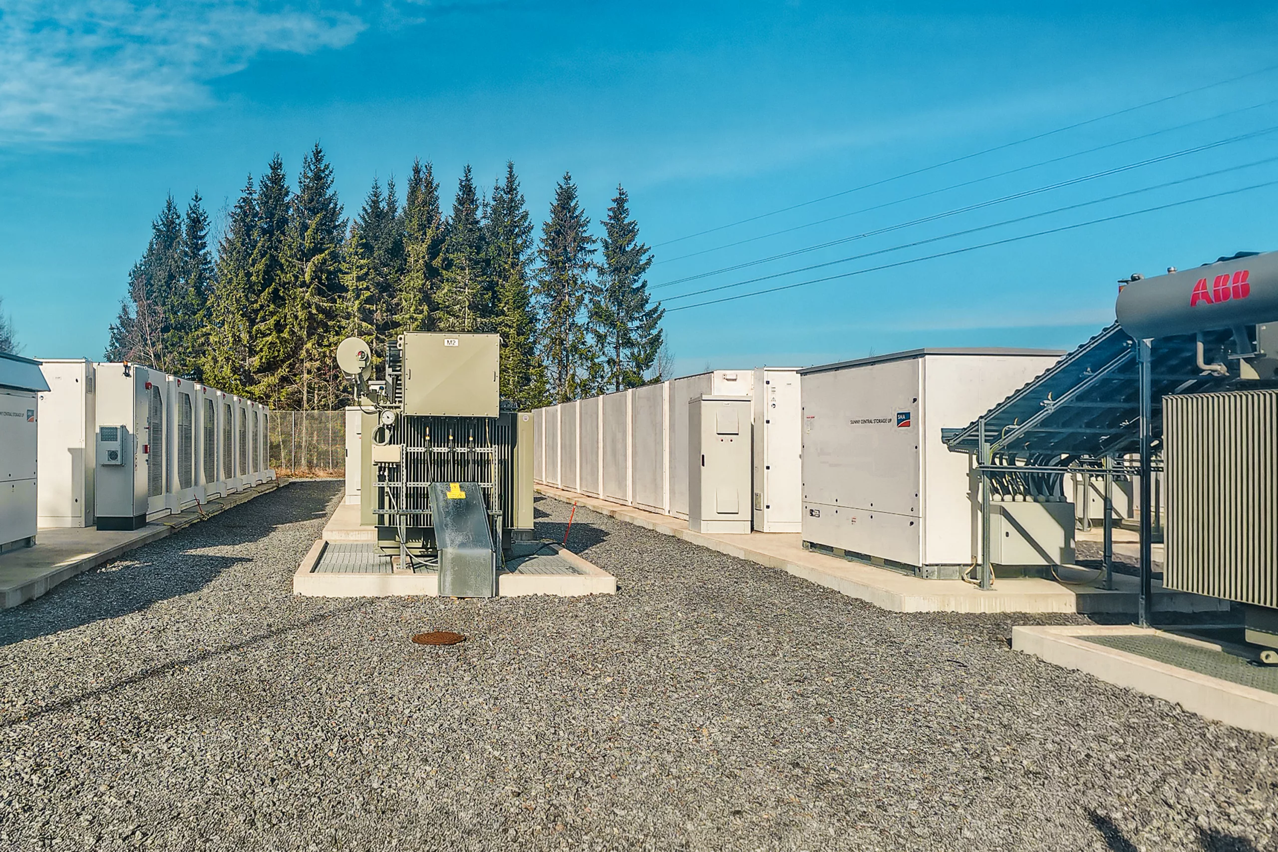 Multiple white battery storage units with cooling systems and control panels arranged in rows, photographed during golden hour with backlit sky