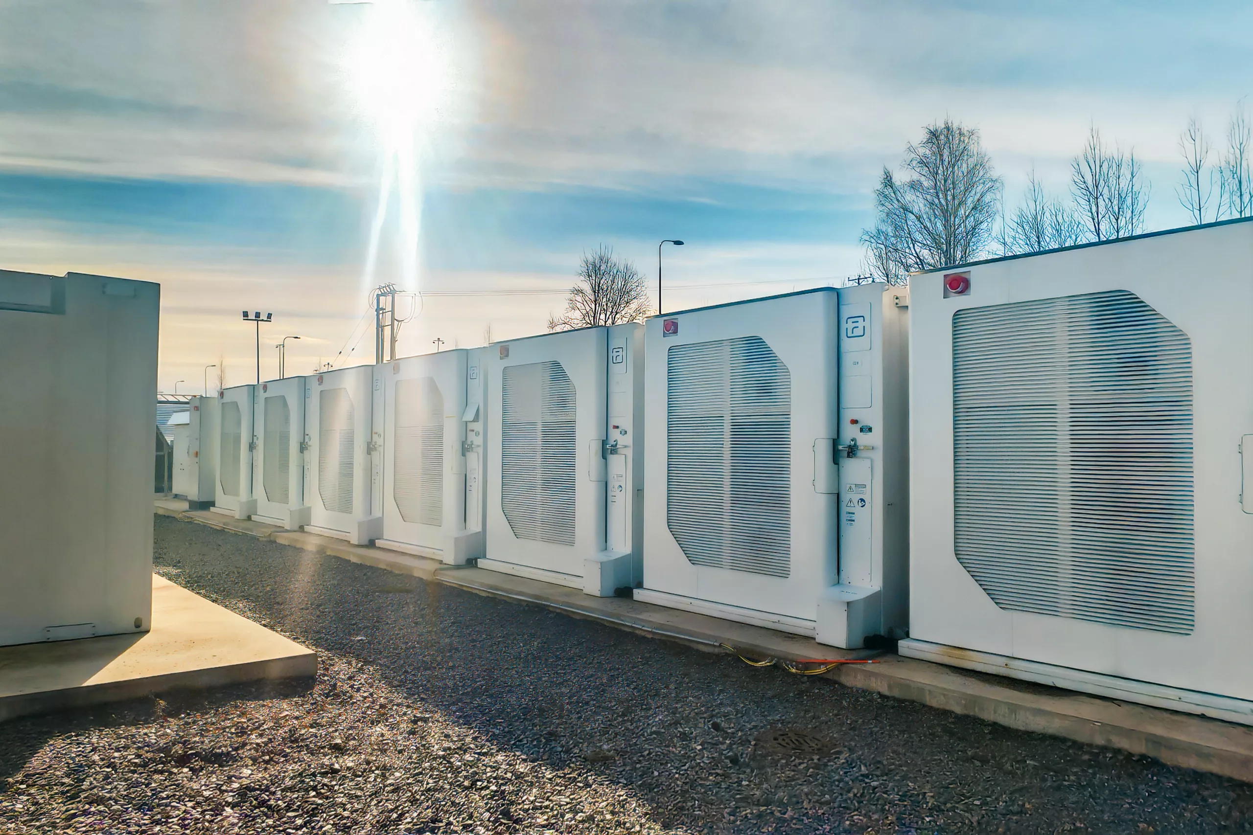 Multiple white battery storage units with cooling systems and control panels arranged in rows, photographed during golden hour with backlit sky