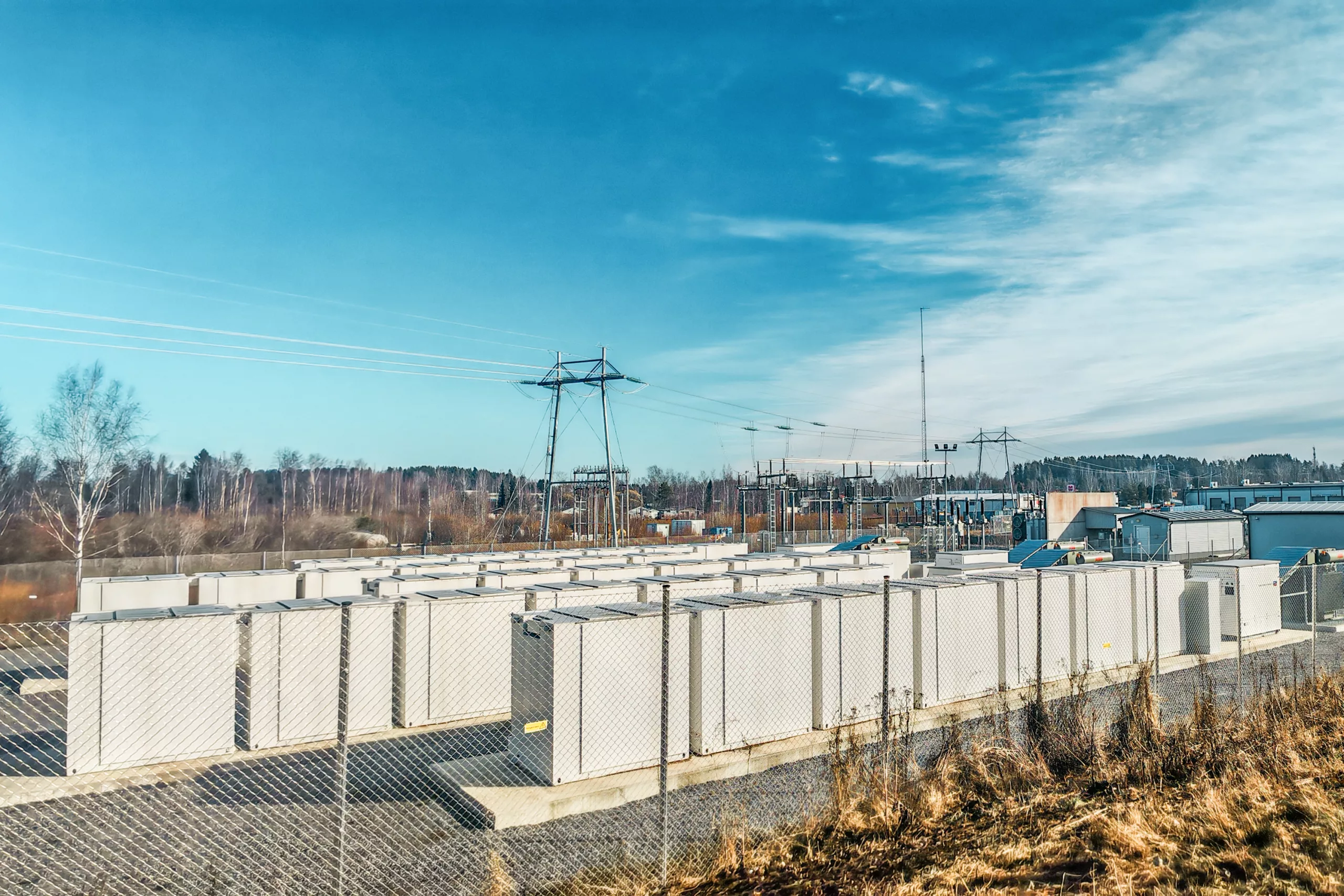 The battery storage field displays an ABB transformer station, white storage modules, a high-voltage transmission line, and a tank storage system set against a backdrop of coniferous trees.