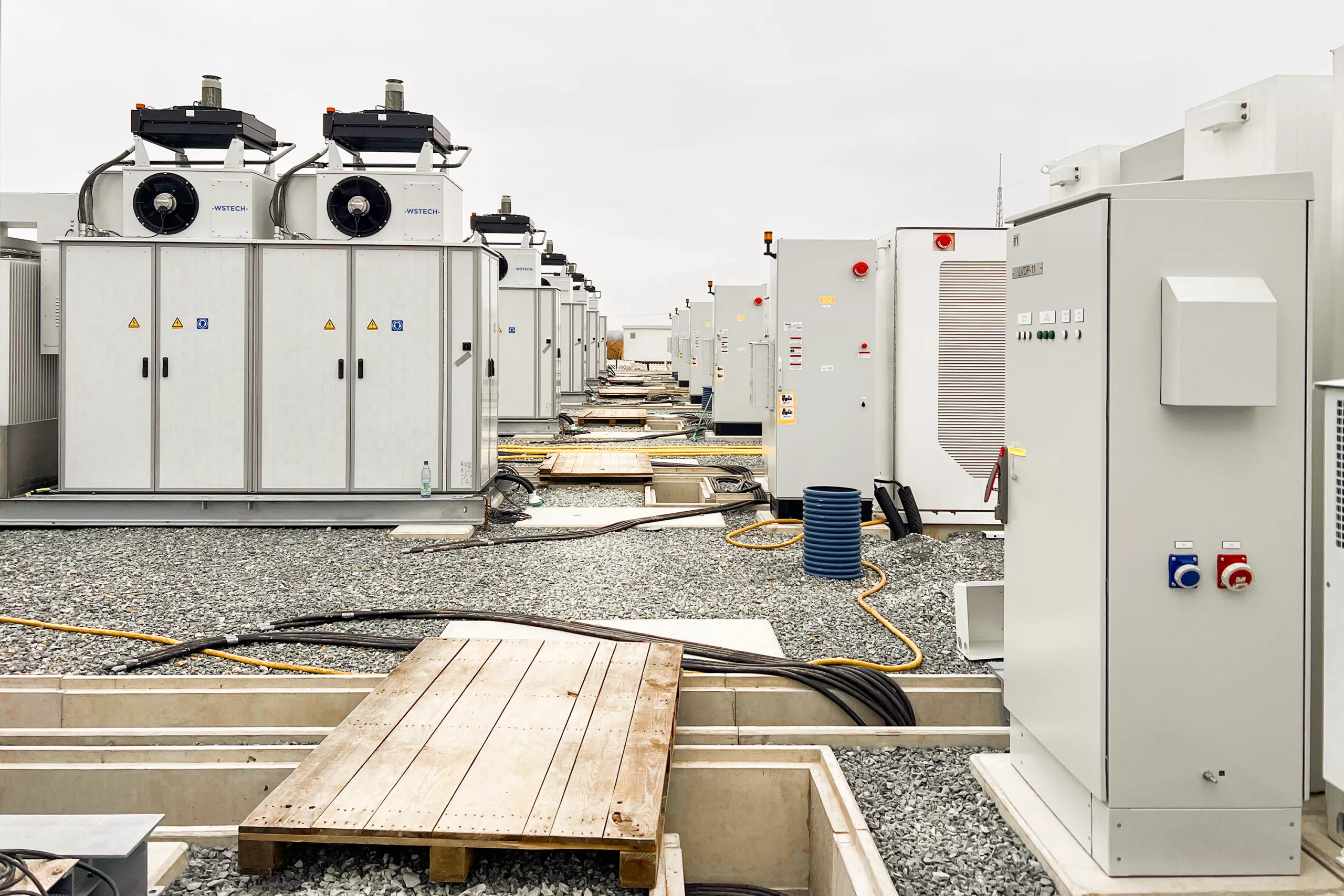 Rows of battery storage units with switchgear cabinets and cooling systems on a gravel surface at a utility-scale BESS site under construction.