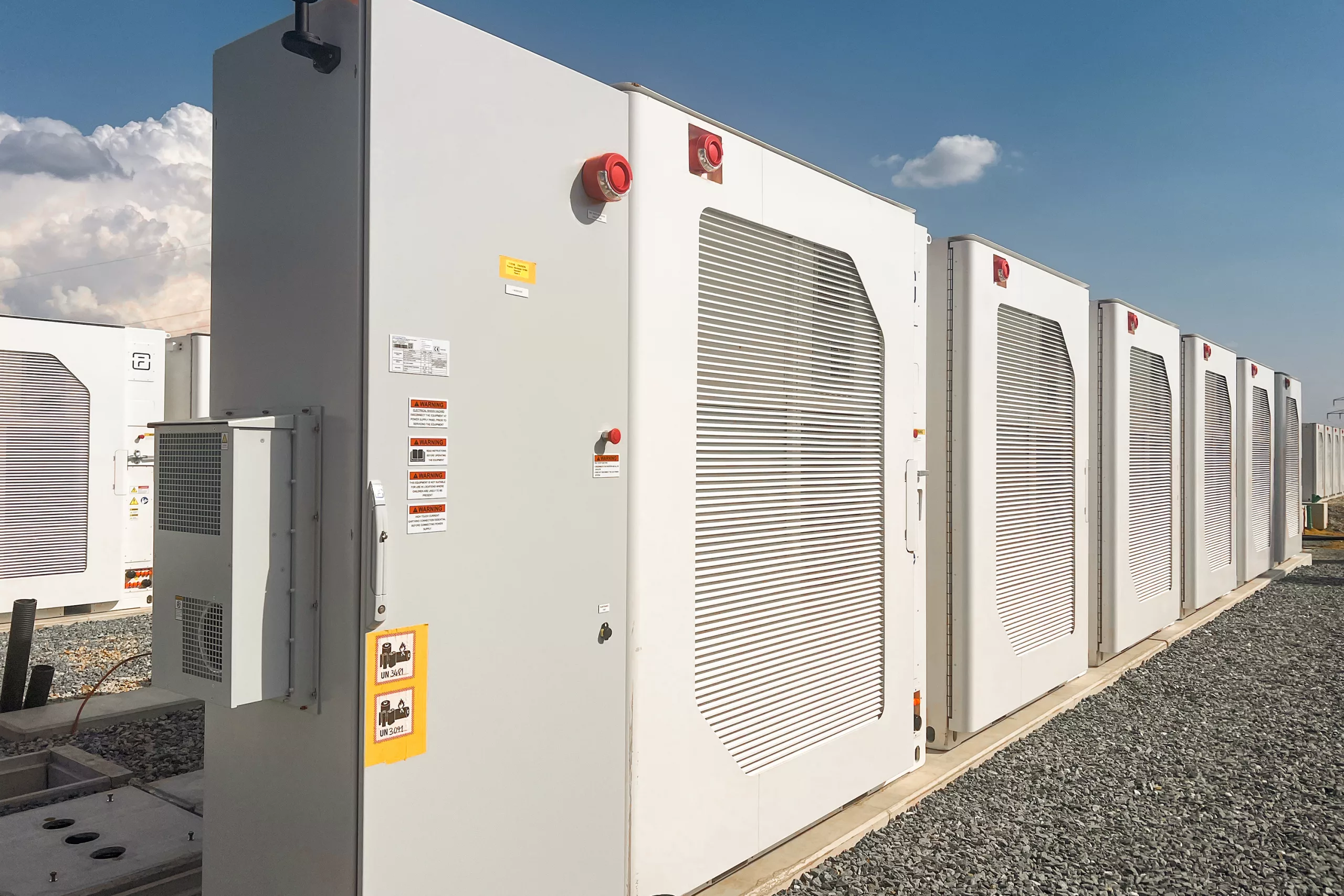 Exterior view of multiple large-scale battery energy storage containers featuring ventilation grilles, emergency shut-off switches and hazard labels.