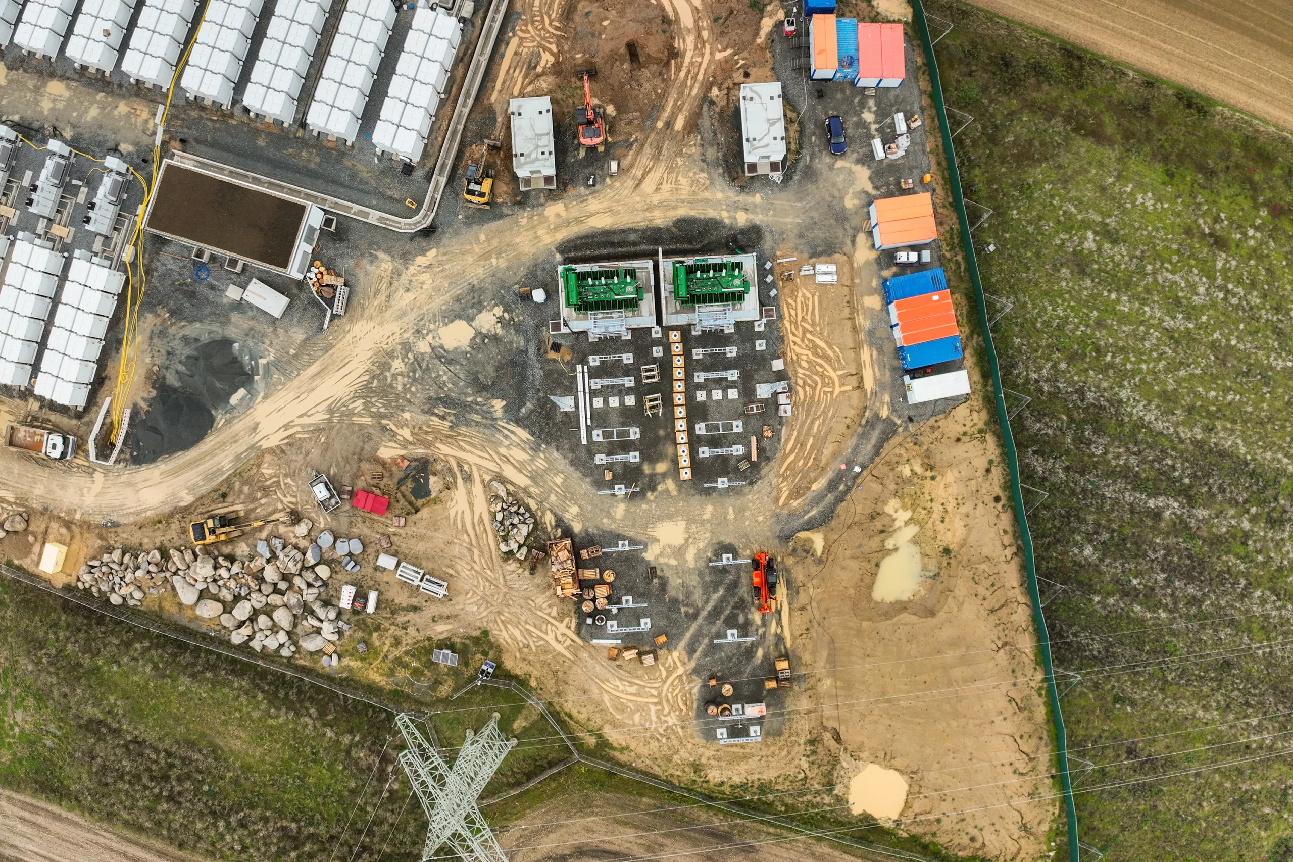 Aerial view of a utility-scale battery energy storage facility under construction, showing installed storage containers, prepared foundations, construction vehicles, transformers and a high-voltage pylon at the edge of the site.