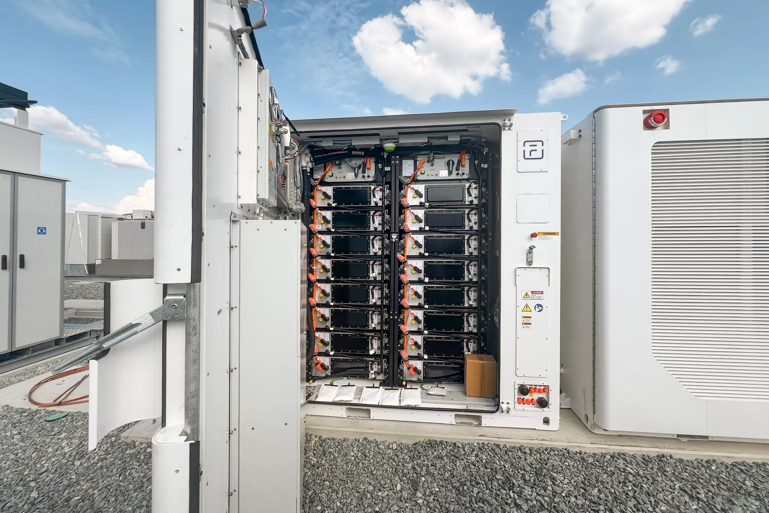 Opened battery energy storage container revealing stacked lithium-ion battery modules with orange high-voltage connectors and internal wiring at an outdoor utility-scale storage facility.