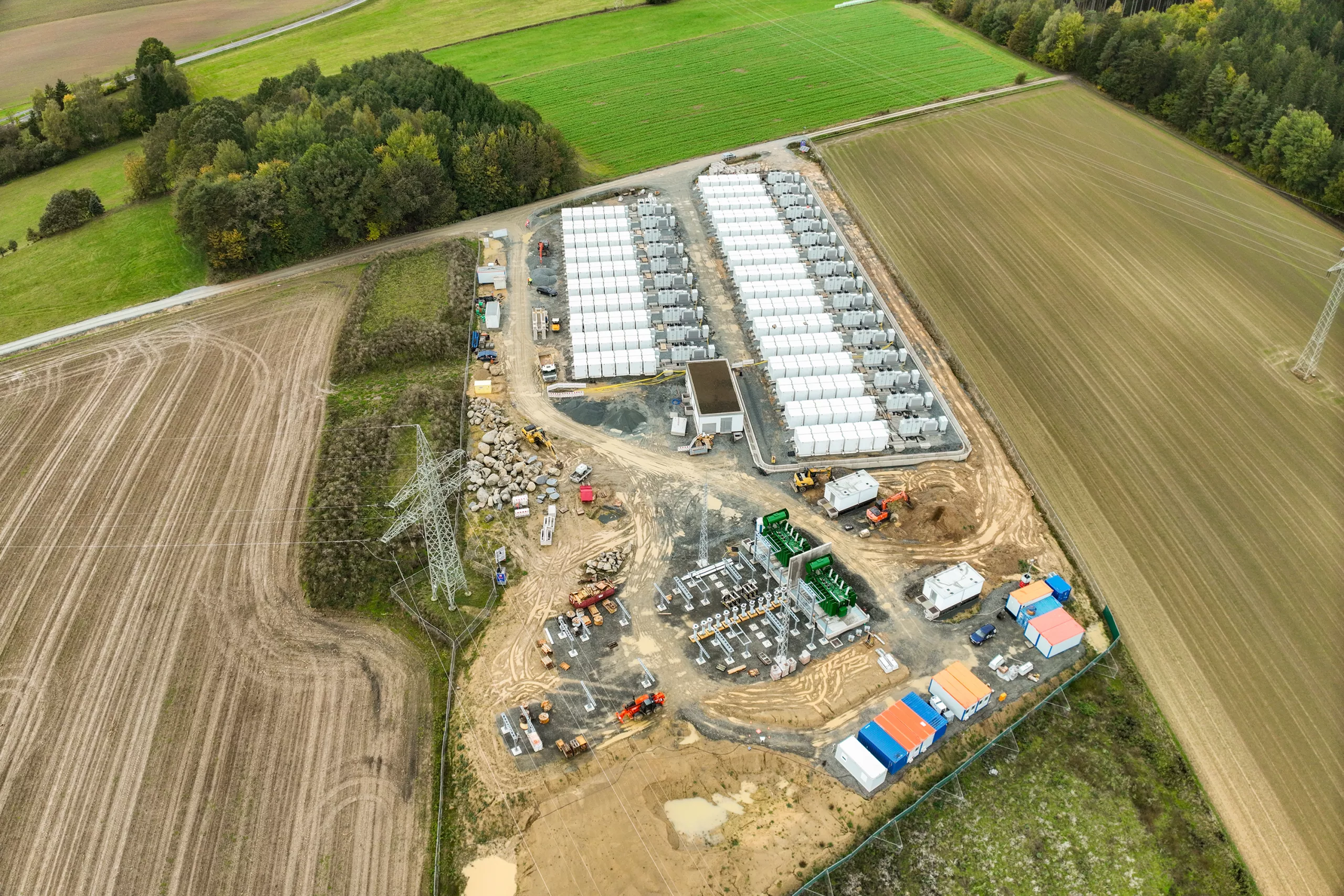 Drone shot of a utility-scale battery energy storage facility with multiple rows of installed storage containers, a substation under construction and high-voltage pylons, surrounded by agricultural fields.