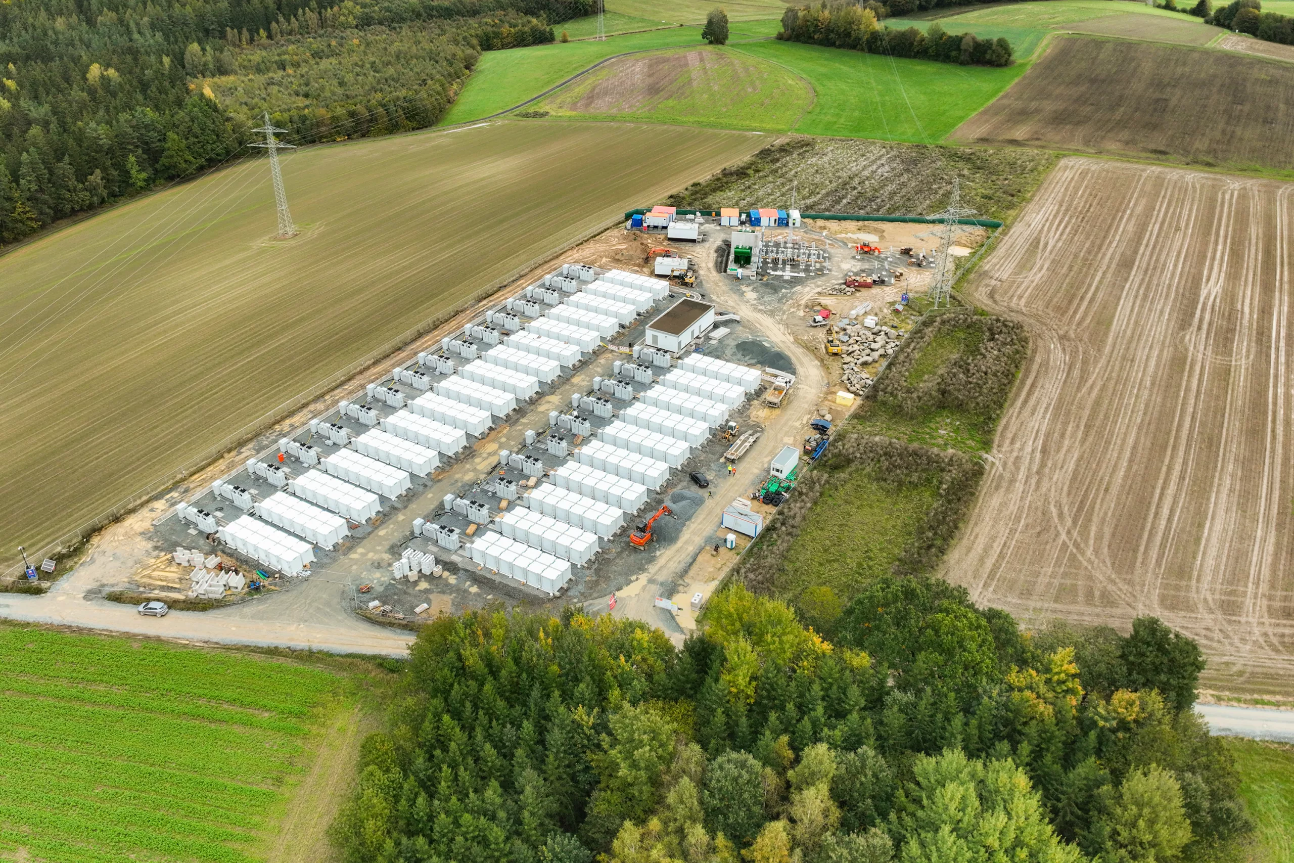 Drone shot of a utility-scale battery energy storage facility under construction with densely arranged storage containers, a substation and construction vehicles, surrounded by agricultural fields and woodland edges.