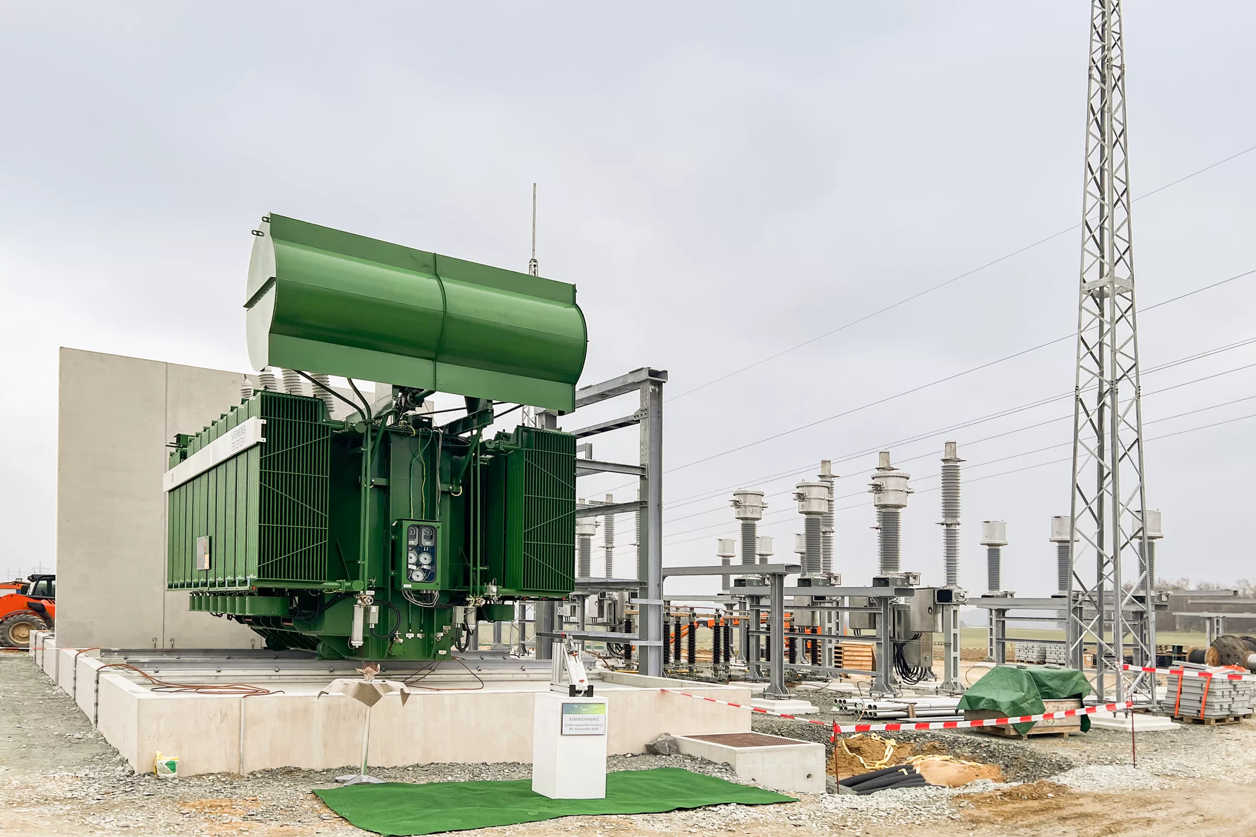 Large power transformer with oil cooler on a concrete foundation next to a high-voltage switchgear with insulators and overhead line masts at a substation under construction.