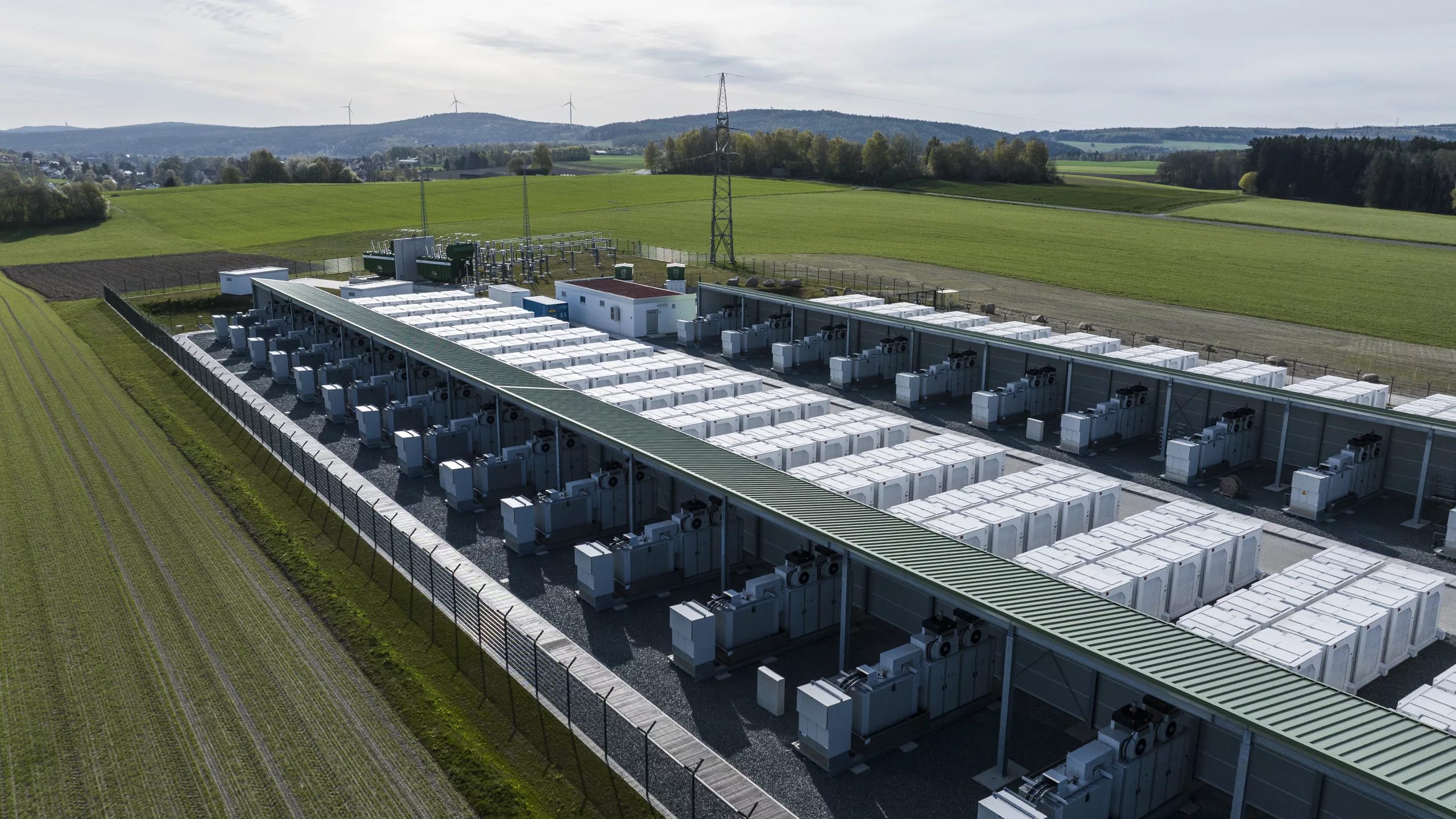 Oblique aerial view of a battery storage plant showing rows of battery cabinets and inverters under open canopy structures, with transformer substation and wind turbines in the background.