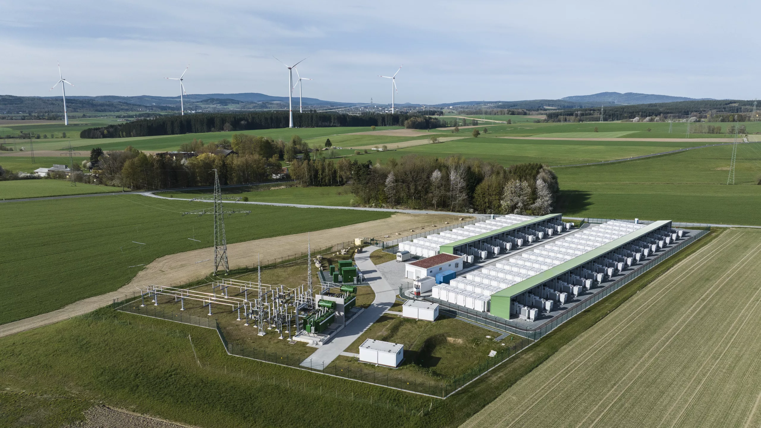 Aerial view of a large-scale battery energy storage system (BESS) with transformer substation in a rural landscape, wind turbines and high-voltage pylons visible in the background.