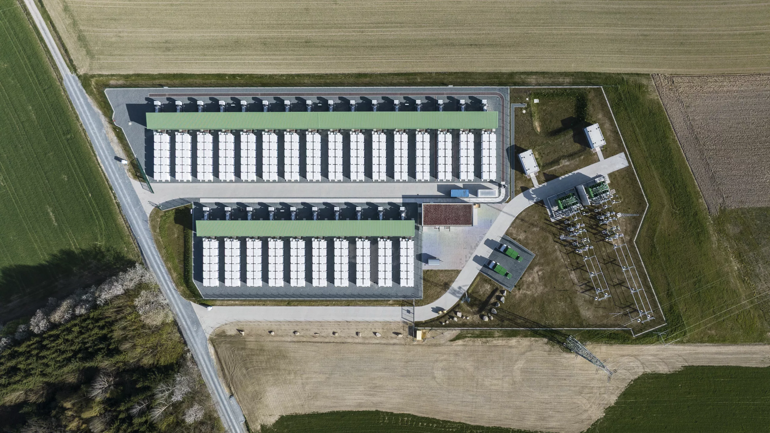 Top-down aerial view of a battery storage facility with two parallel battery block arrays under canopy structures, adjacent transformer substation and access road.