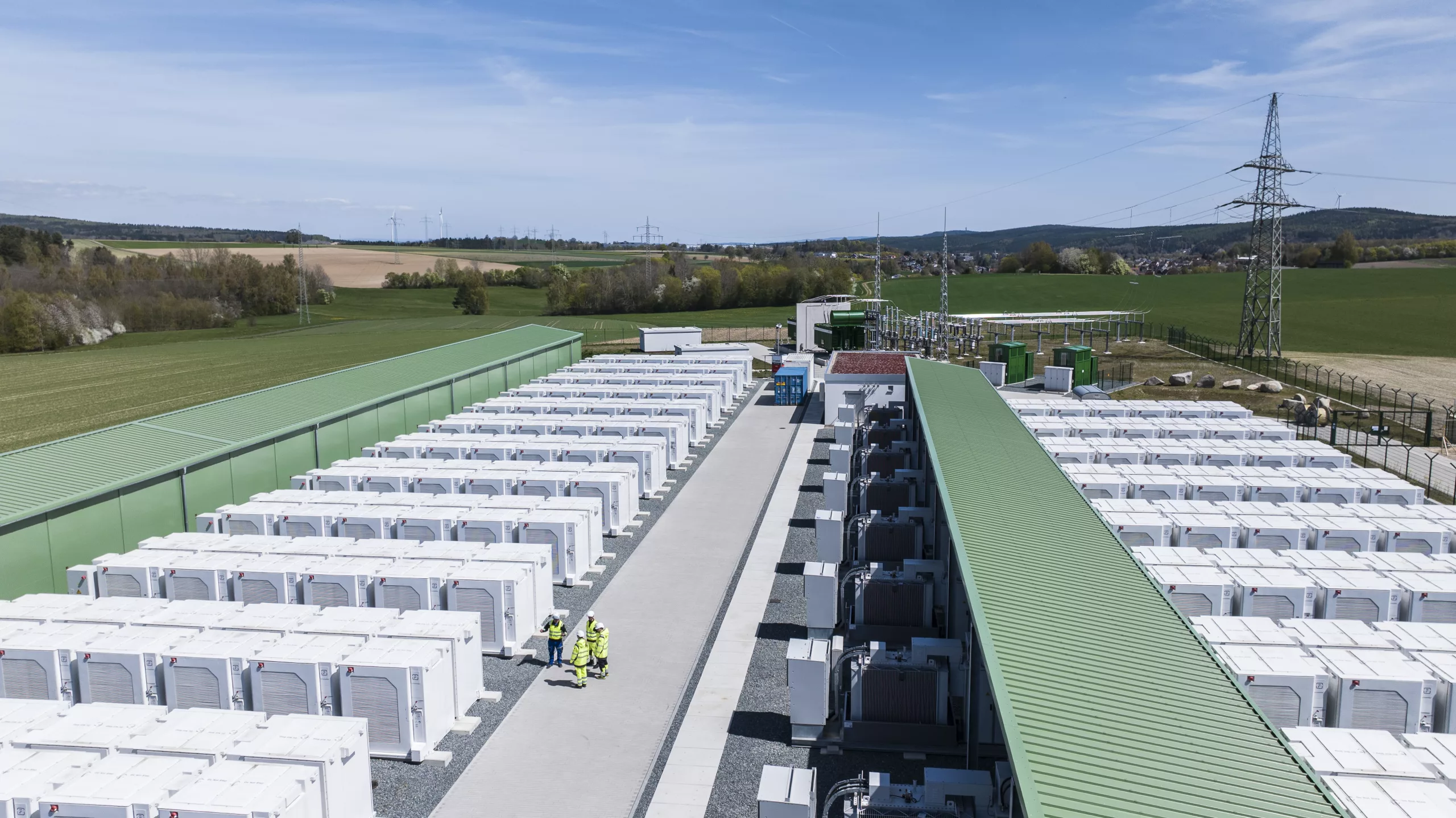 Central walkway of a battery energy storage system with battery cabinets on both sides under green canopy roofs, three engineers in high-visibility vests conducting an inspection, transformer substation visible in the background.